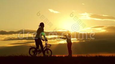 剪影自行车可爱的家庭在草地上日落时间。母亲和婴儿在日落时骑自行车的剪影。生活方式